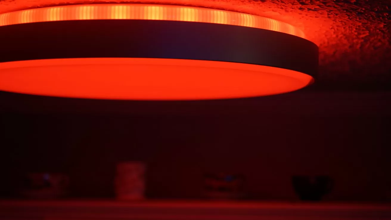 Circular ceiling light glowing bright red against a dark textured ceiling, casting warm red illumination over a dimly lit room with faint objects visible on a shelf below