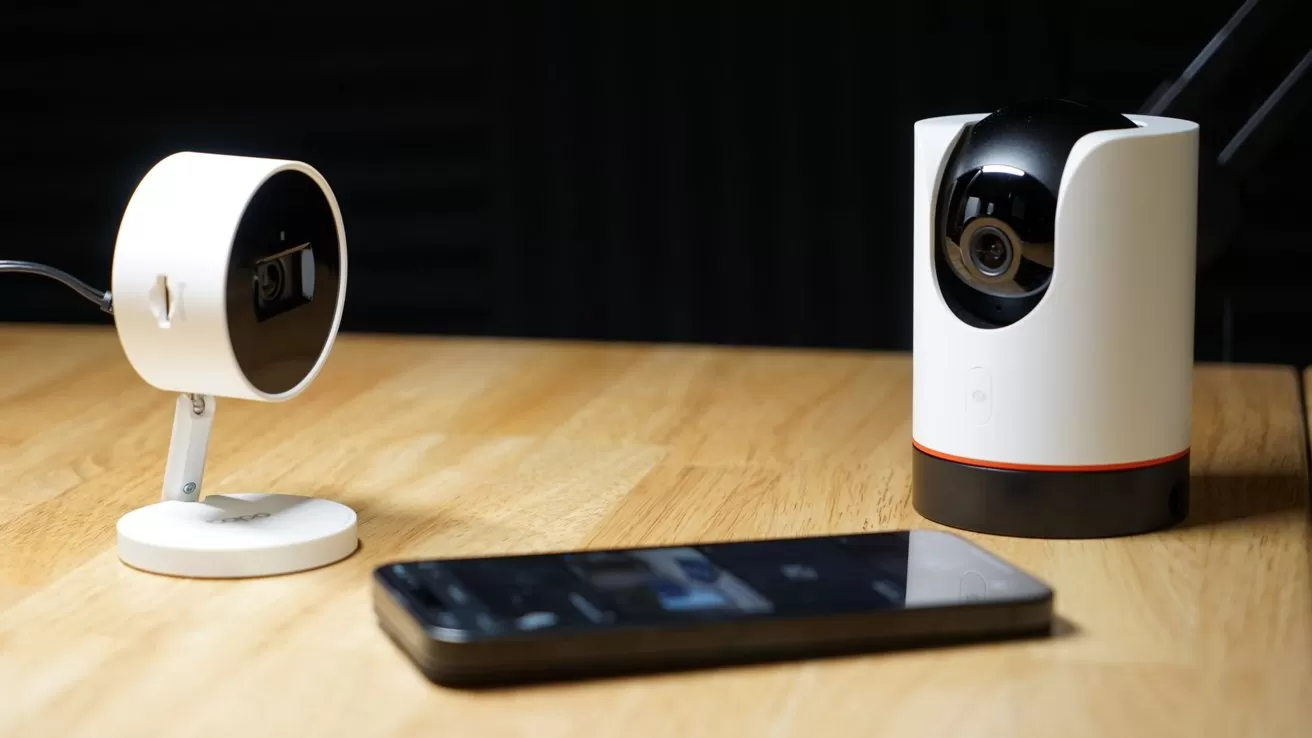Two modern security cameras and a smartphone resting on a wooden table in a dimly lit room, suggesting a smart home surveillance setup