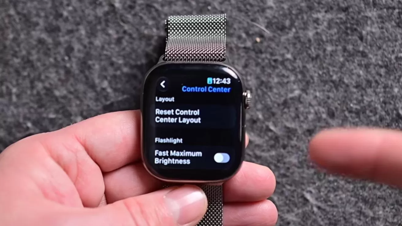 Hand holding an Apple Watch with metal band, screen showing Control Center settings menu, while another finger points toward the watch against a gray textured background