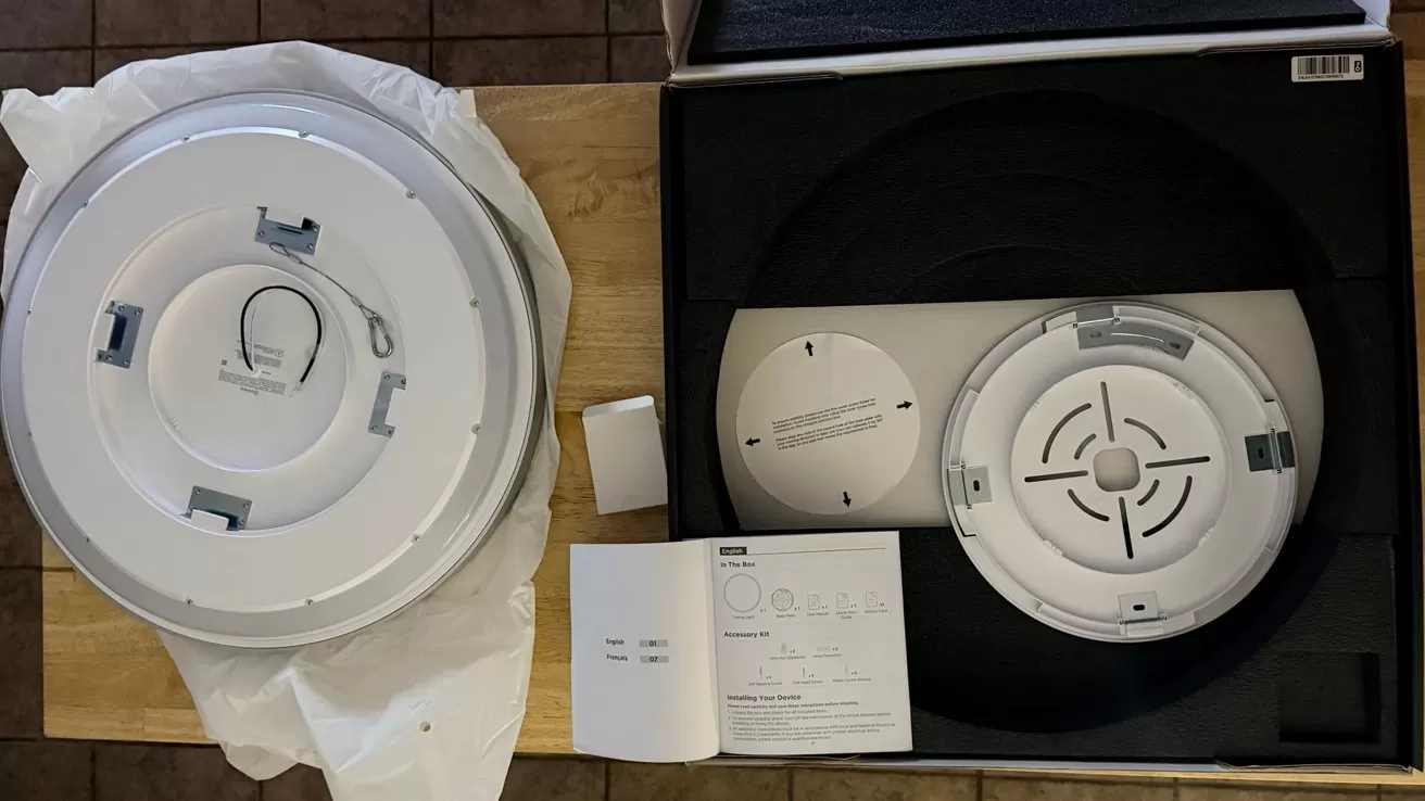 Ceiling light fixture unboxed on a table, with one round light lying on a cloth and another still in packaging, alongside mounting hardware and instruction manual