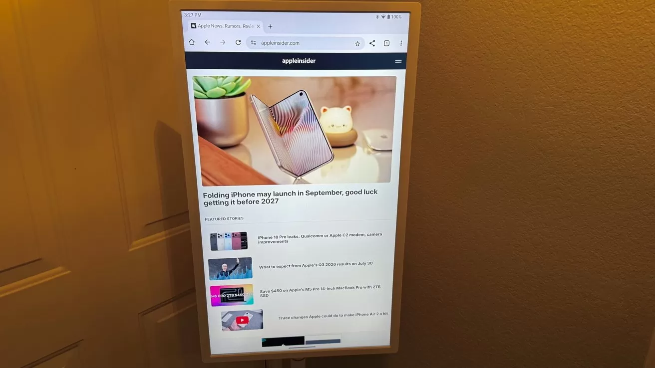 Vertical computer monitor mounted on a wall displaying a tech news website about folding iPhones, with photos, article titles, and a soft yellow room light in the background