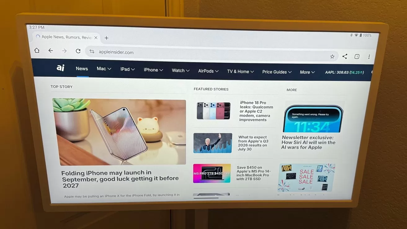 Computer monitor mounted on a wall displaying a technology news website, featuring articles about iPhones and Apple products with headlines, thumbnails, and navigation menu in a bright web browser interface