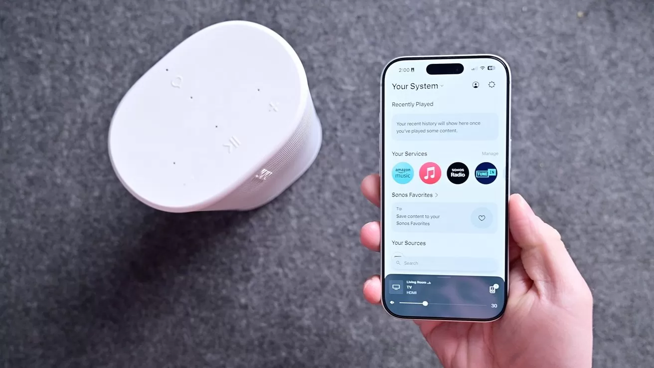 Hand holding a smartphone with a home audio app open, next to a white wireless speaker on a gray surface