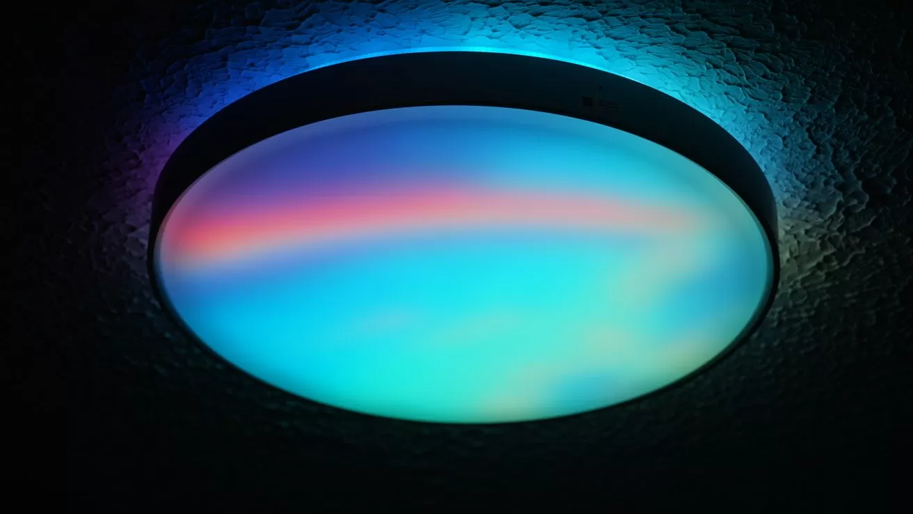 Round ceiling light glowing with soft gradient colors of blue, cyan, and pink, casting a faint halo on the textured ceiling in a dark room