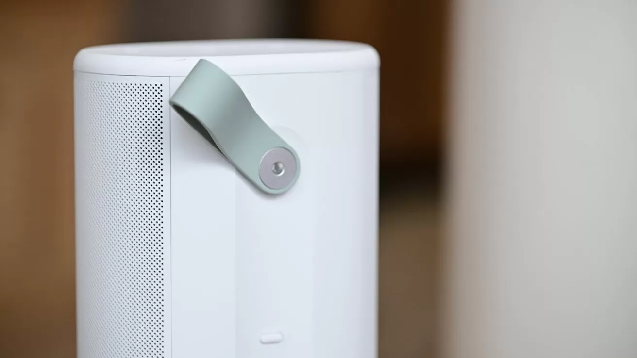Close-up of a modern white portable speaker with side perforated grille and a light gray carrying strap attached by a metal button, set against a softly blurred indoor background