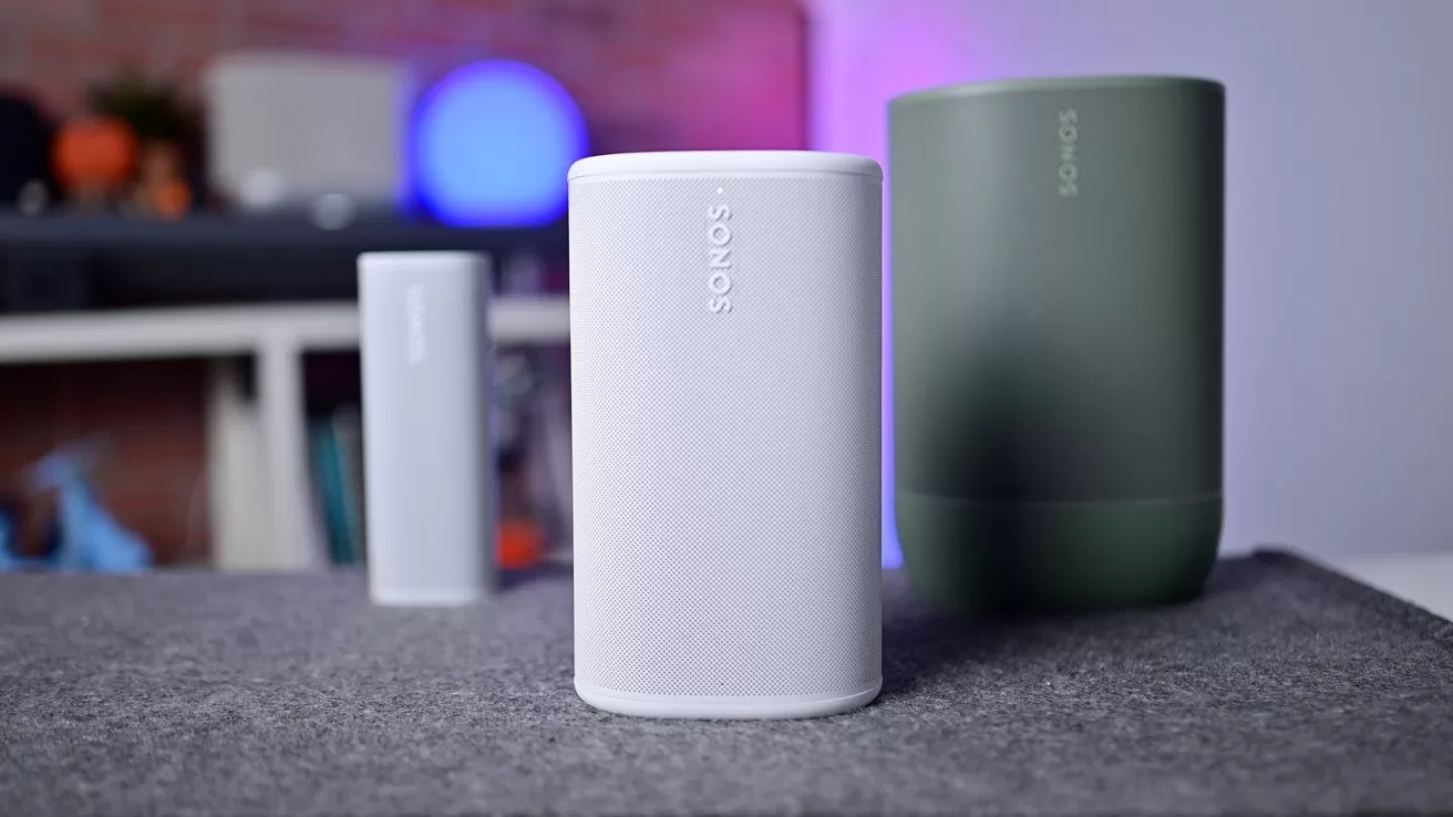 Three modern Sonos speakers on a gray surface, with a white cylindrical speaker in front, a larger green speaker to the right, and a smaller white speaker blurred in the background