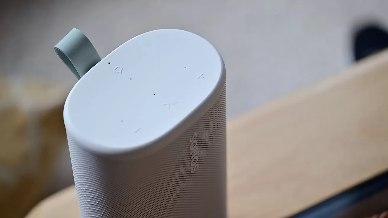 Close-up of a white cylindrical Sonos speaker on a wooden surface, showing touch controls on top and a small loop handle, with a softly blurred indoor background