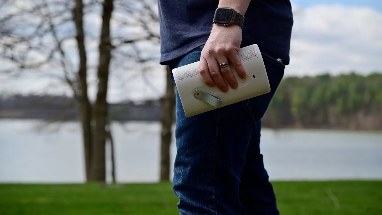 Person standing outdoors near a lake and trees, wearing casual clothes and a smartwatch, holding a white portable speaker or device by their side on a grassy area