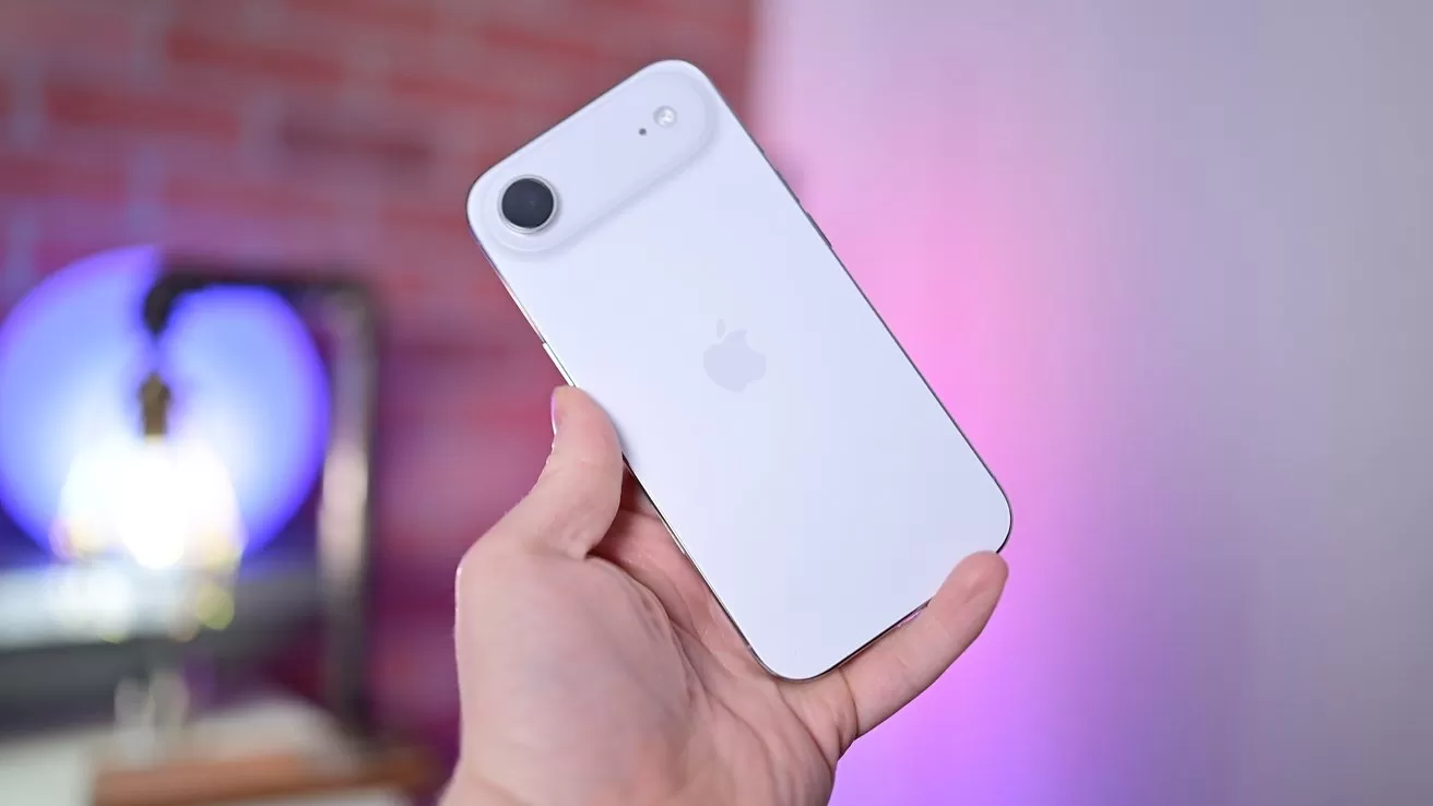 Hand holding a white iPhone, showing the back with Apple logo and single rear camera, against a softly blurred background with pink and purple lighting and a brick wall.