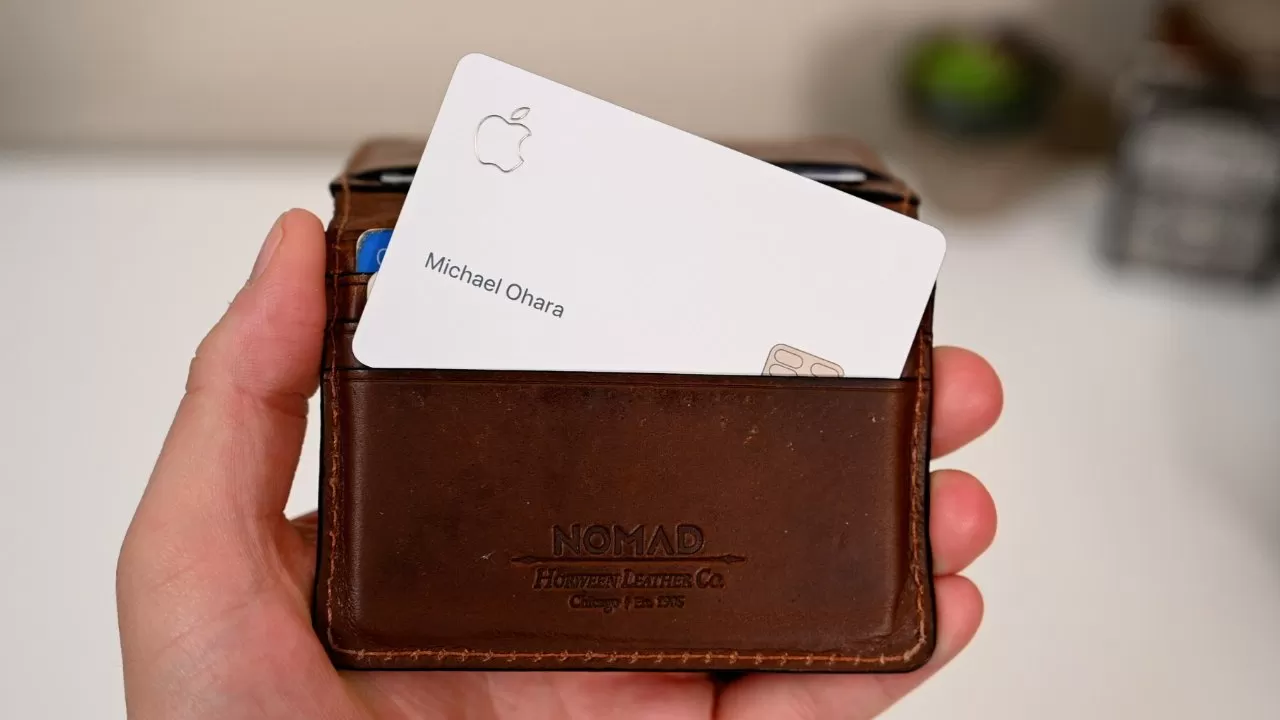 Hand holding a brown leather NOMAD wallet with a white Apple credit card labeled Michael Ohara partially visible in the front slot, against a blurred indoor background