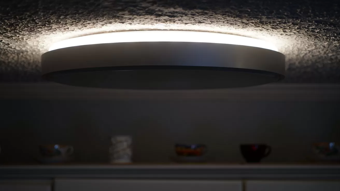 Circular ceiling light glowing softly against textured ceiling, casting halo of light; below, dimly lit shelf displays several coffee cups spaced along the top of kitchen cabinets