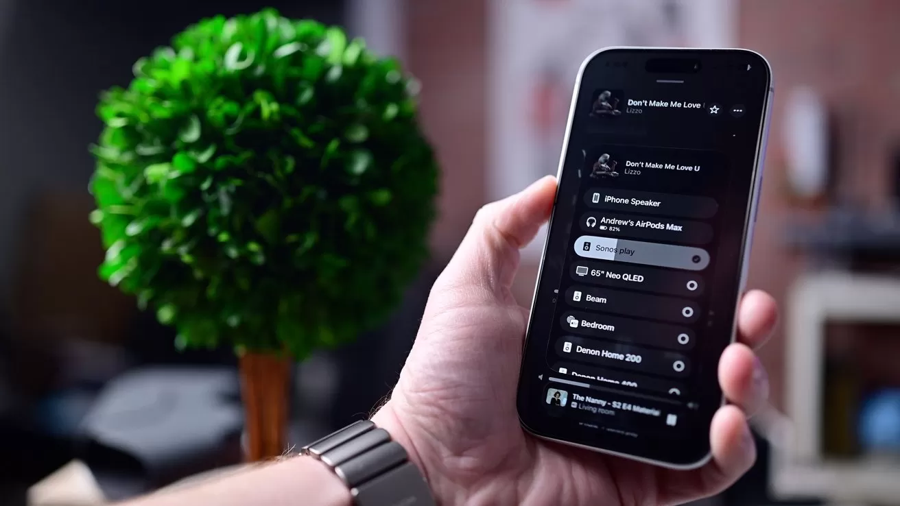 Hand holding a smartphone displaying a dark-mode playlist or music app interface, with a blurred green potted plant and home interior in the background