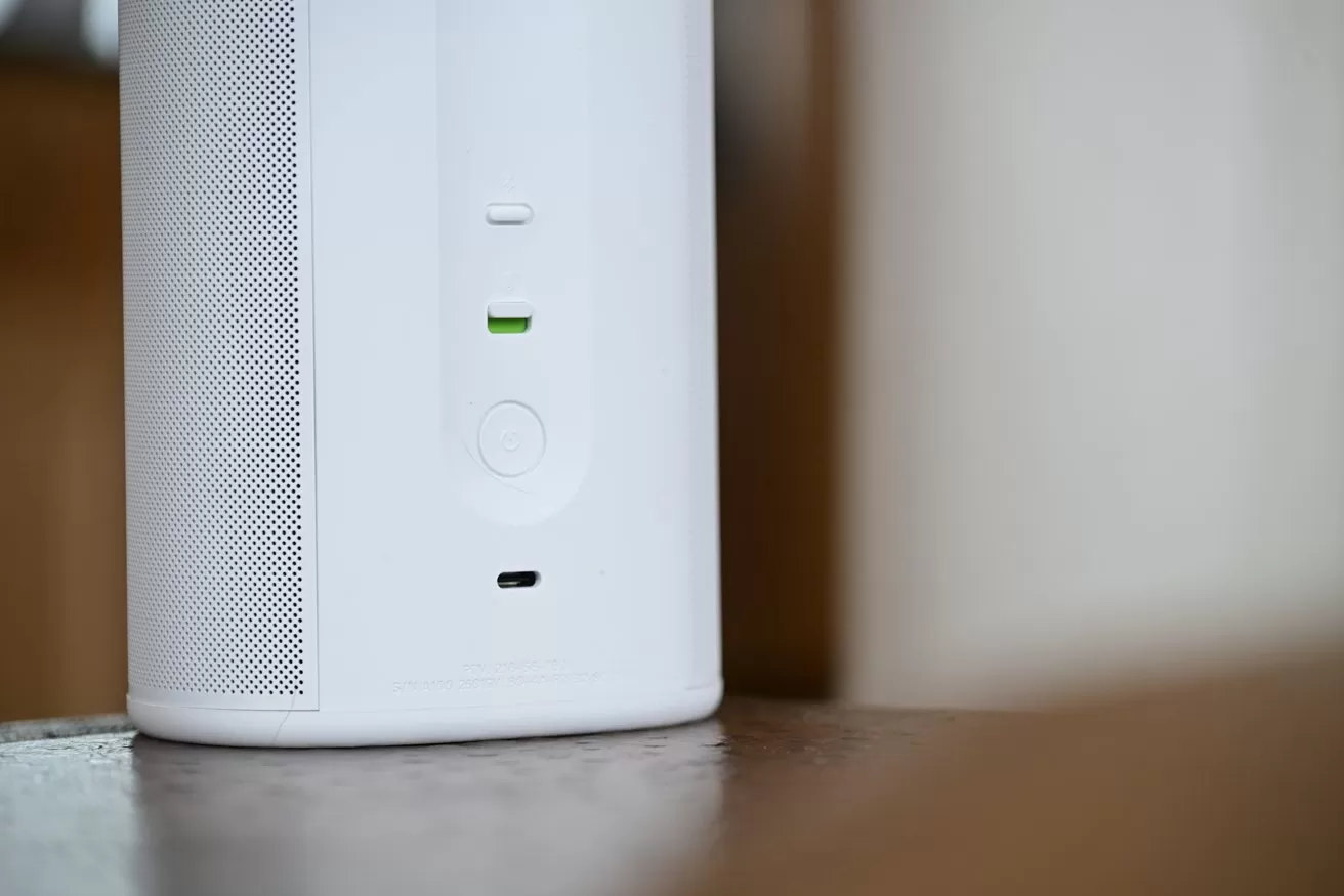 Close-up of a modern white smart speaker showing mesh side, small LED indicators, a round button, and a charging port, sitting on a wooden surface indoors