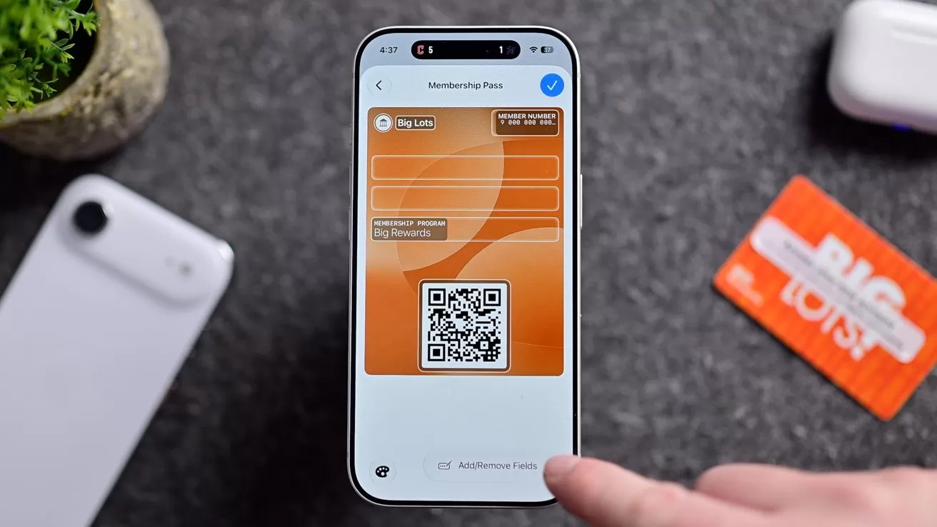 Smartphone displaying a digital membership card with QR code on screen, surrounded by a physical membership card, another phone, earbuds case, and small potted plant on a gray surface
