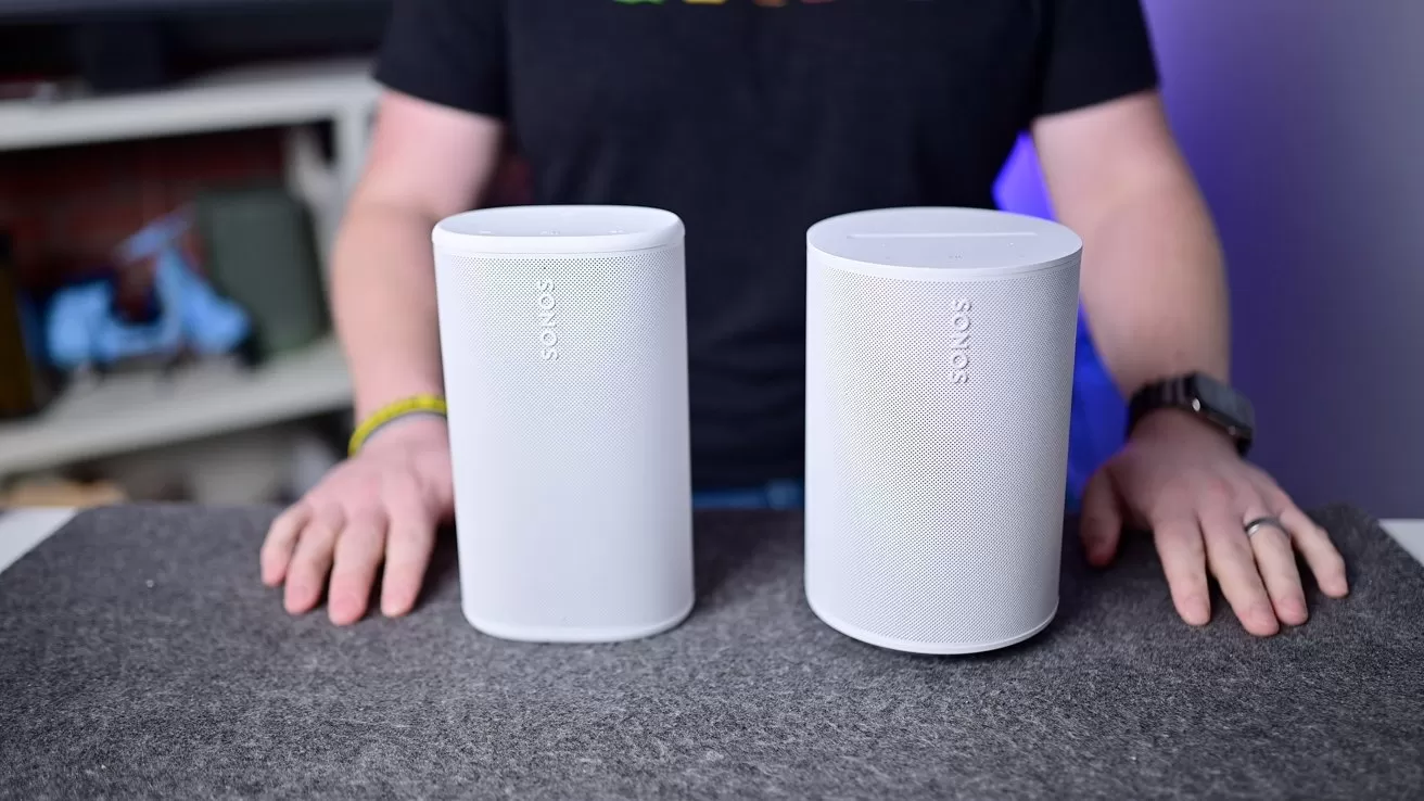 Person standing behind a table with two white Sonos smart speakers placed side by side on a gray surface, hands resting near each speaker, blurred shelves and lights in the background