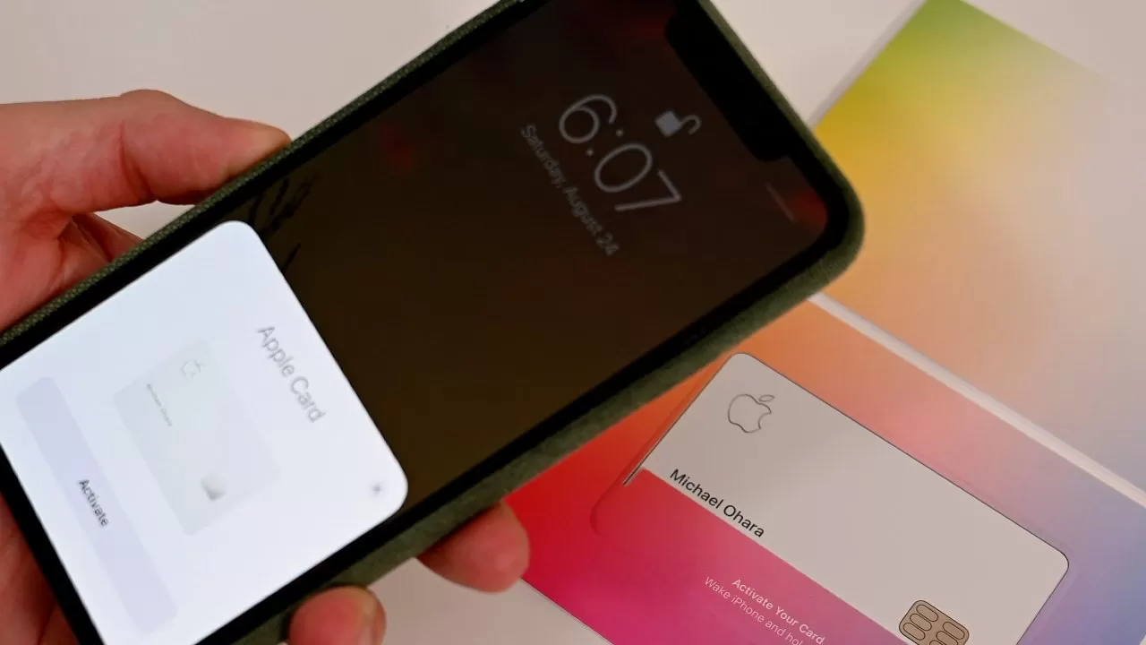 Hand holding an iPhone displaying Apple Card activation screen near a physical Apple Card on a colorful gradient holder, showing contactless setup process for the credit card