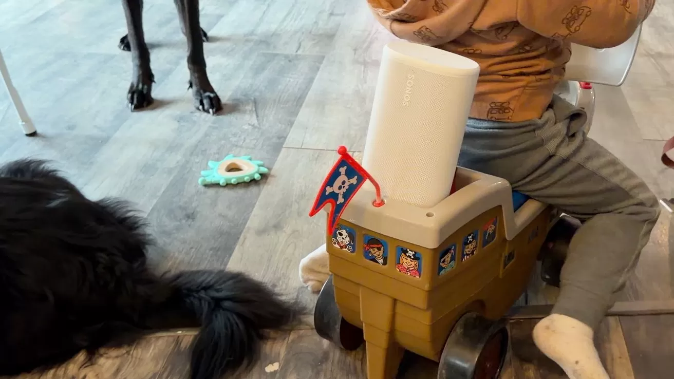 Toddler sitting on a toy pirate ship with wheels indoors, near a large black dog lying on the floor and scattered toys on wooden flooring