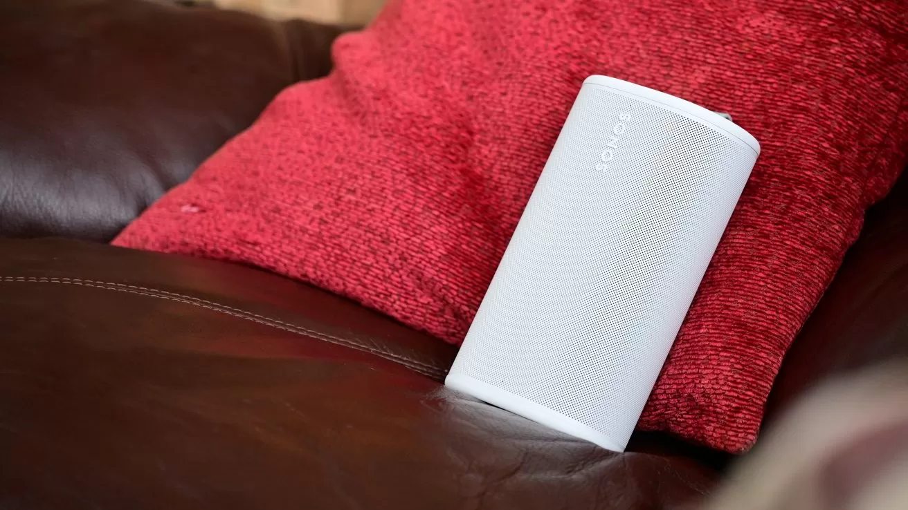White Sonos speaker resting against a bright red cushion on a brown leather sofa, suggesting a cozy living room setting with casual placement of the speaker