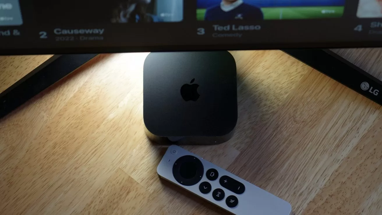 Small Apple TV box beneath a television on a wooden table, with a slim white remote control lying in front, screen displaying streaming content at the top edge