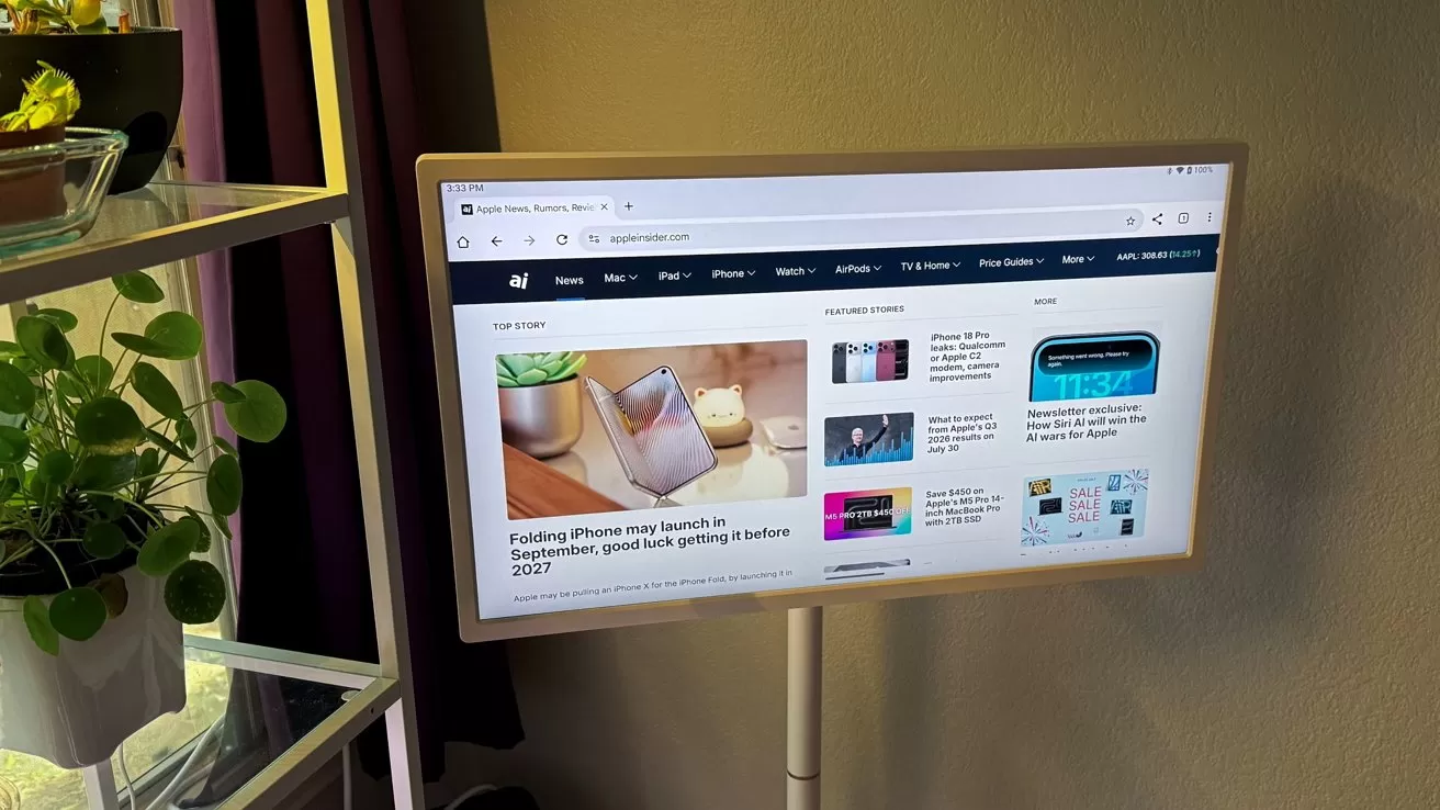 Computer monitor on a tall stand displaying a technology news website, positioned beside a shelving unit with potted plants, against a plain indoor wall with soft lighting