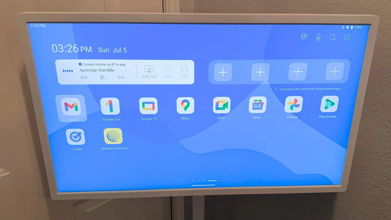 Wall mounted tablet-like screen displaying a blue home screen with time, date, and various app icons including email, calendar, maps, photos, and settings arranged in a grid