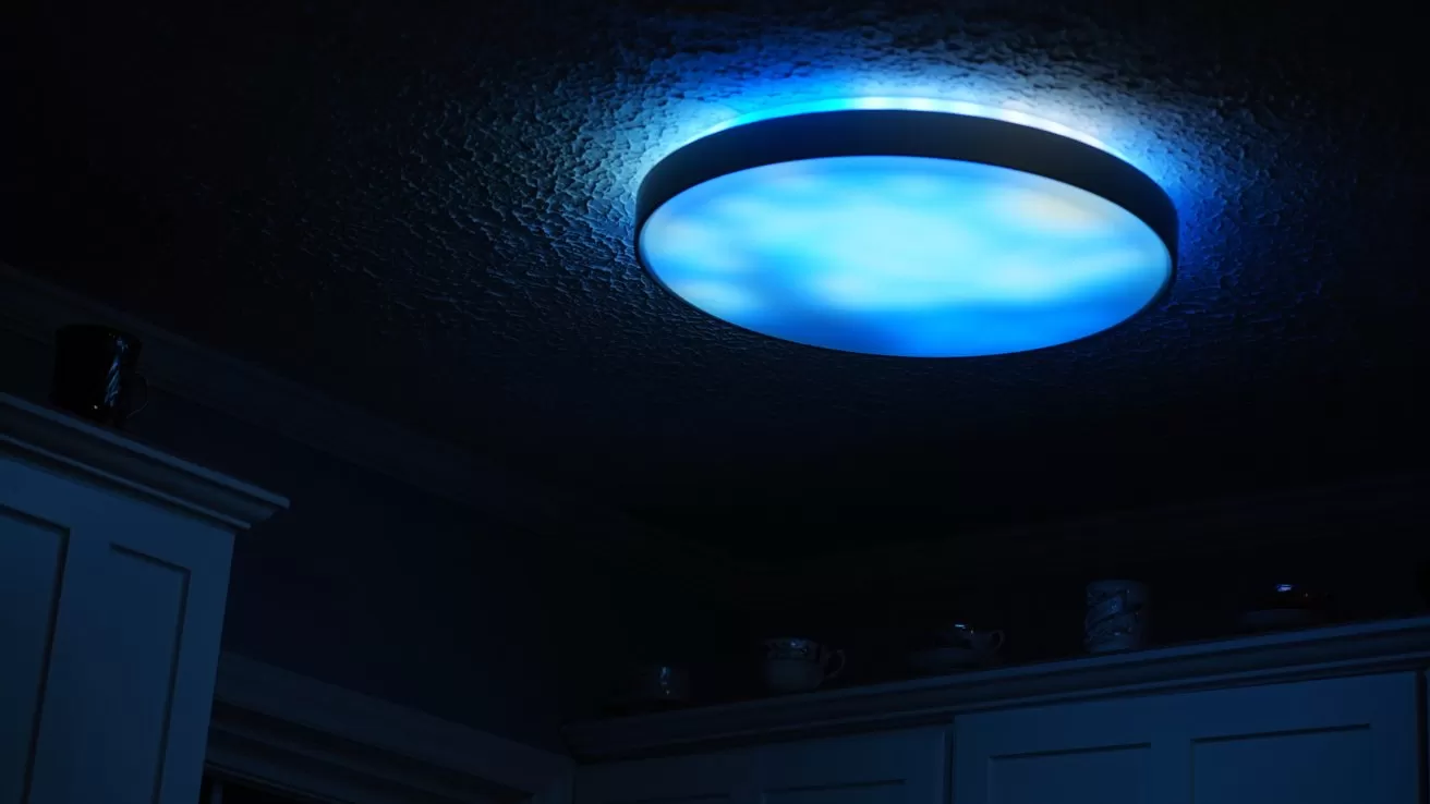 Circular ceiling light glowing with soft blue illumination in a dark room, casting gentle light on textured ceiling and upper kitchen cabinets below